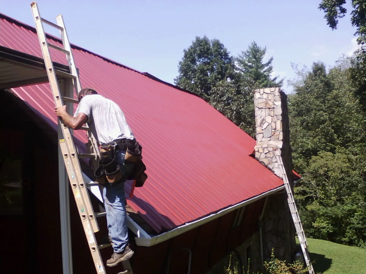 Roofer on a ladder beside a red metal roof and stone chimney — Soffit & Fascia Repair by Roofing Brothers in Carnation